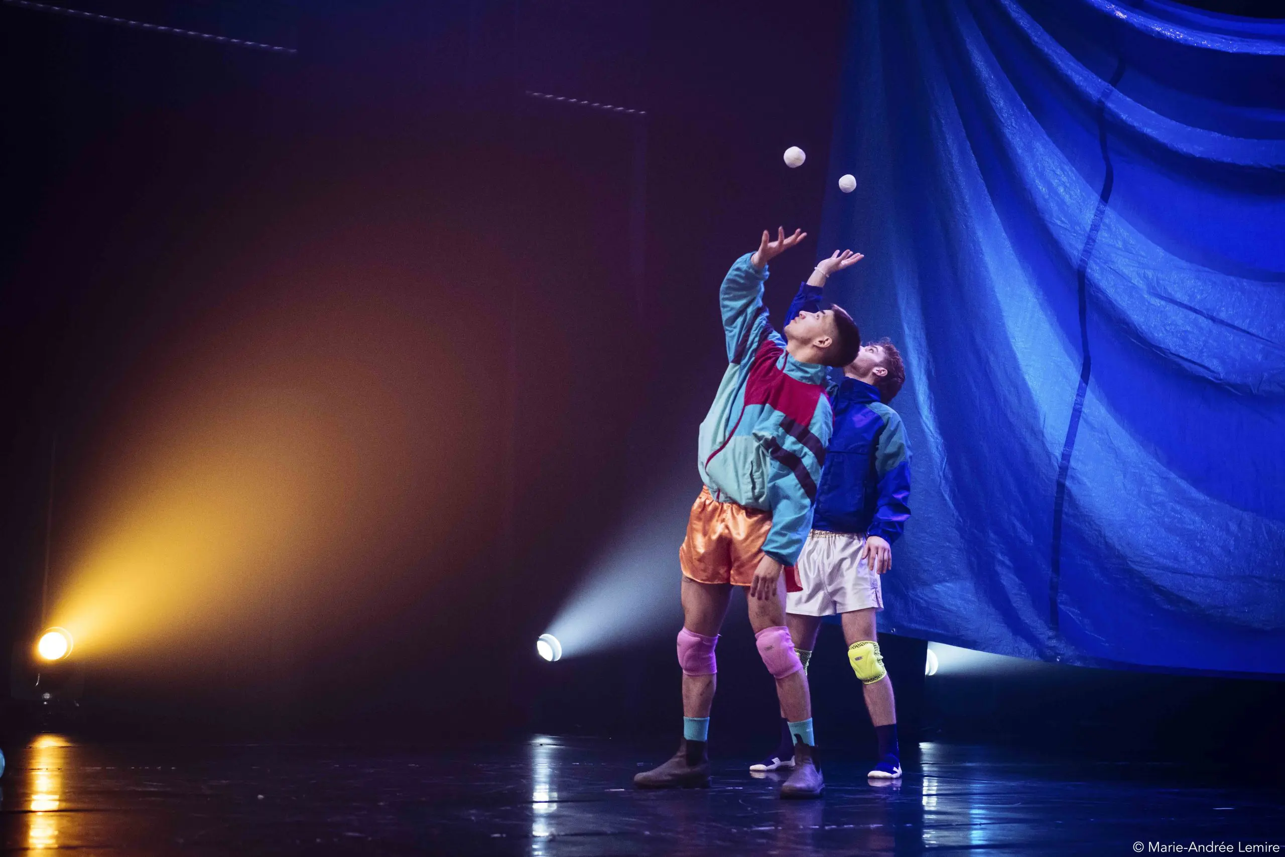 Deux artistes, Zachary Martel et Santiago Rivera Laugerud, vêtus de tenues athlétiques colorées, jonglent avec des balles blanches sur une scène. Un éclairage dramatique et des rideaux bleus plantent le décor alors que l'un d'eux penche la tête en arrière pour équilibrer la balle.