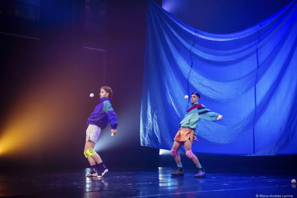 Zachary Martel et Santiago Rivera Laugerud, vêtus de vêtements de sport colorés, jonglent avec des balles sur scène, prenant des poses dynamiques. Une grande toile de fond bleue et un éclairage dramatique créent une atmosphère théâtrale.