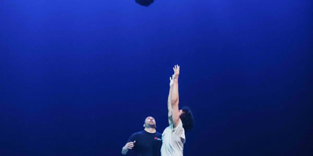 Trois artistes se trouvent sur une scène éclairée en bleu. Oliver Layher est en l'air, la tête en bas dans un flip, tandis qu'une autre personne tend les bras vers lui. Une troisième personne se tient derrière, regardant l'action se dérouler.