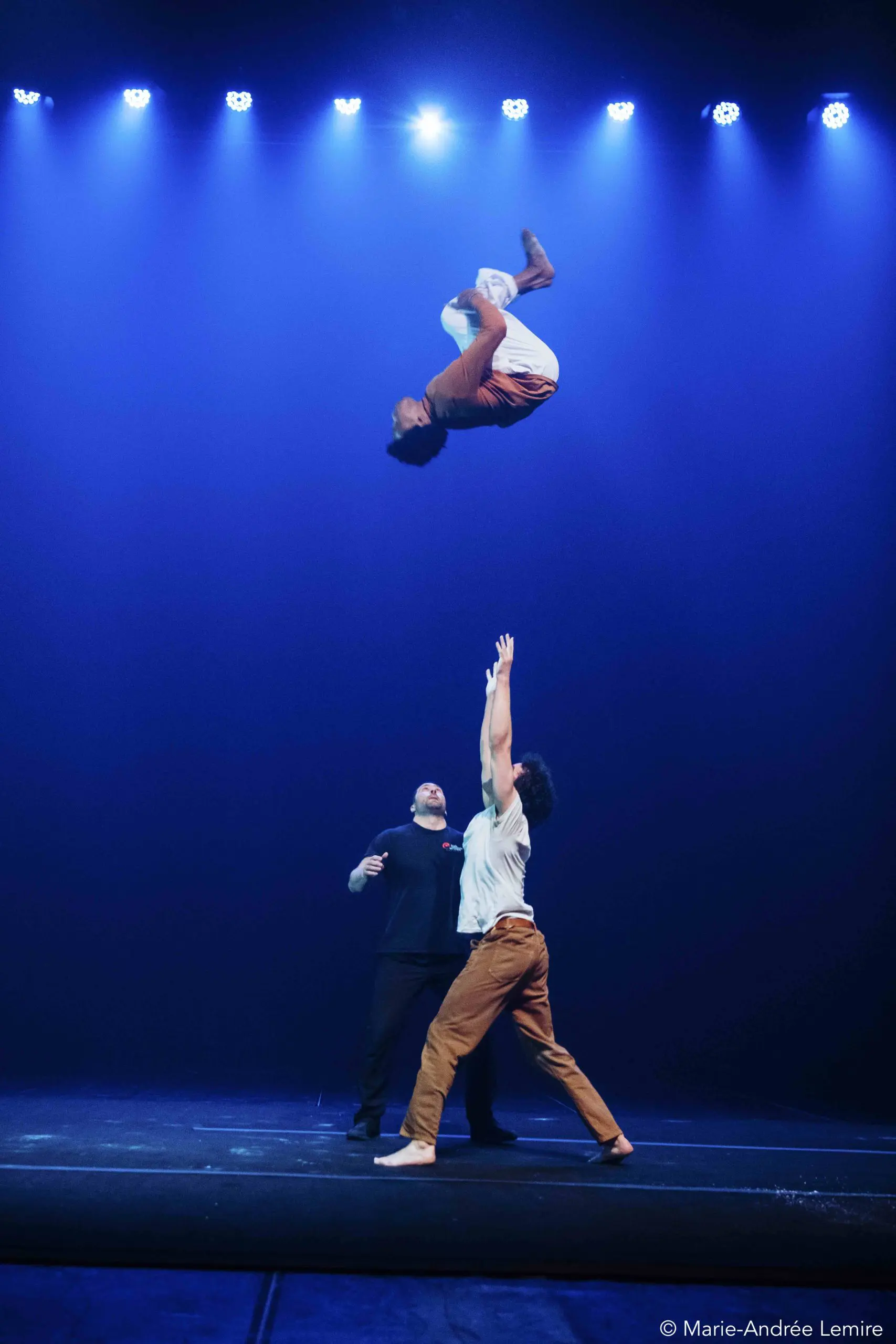 Trois artistes se trouvent sur une scène éclairée en bleu. Oliver Layher est en l'air, la tête en bas dans un flip, tandis qu'une autre personne tend les bras vers lui. Une troisième personne se tient derrière, regardant l'action se dérouler.