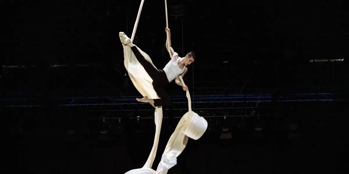 L'artiste aérien Antoine Boissereau, vêtu d'une tenue blanche, pose gracieusement sur des rubans de soie blanche suspendus sur un fond sombre, faisant preuve de force et d'équilibre au-dessus du sol.