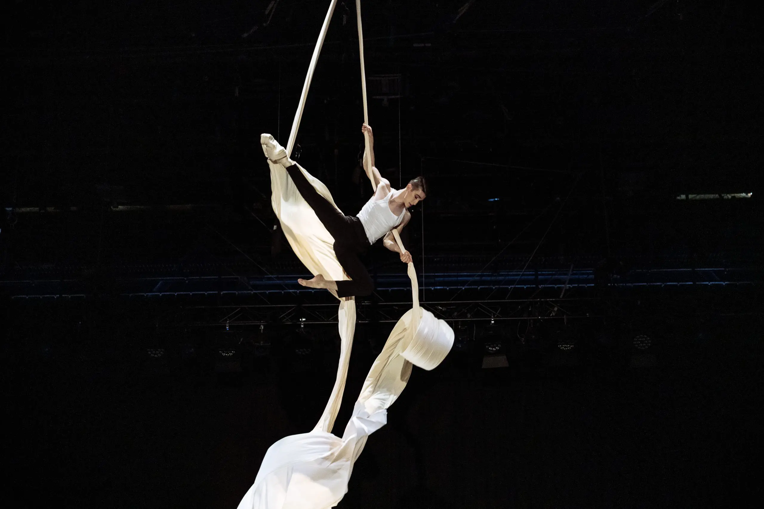 L'artiste aérien Antoine Boissereau, vêtu d'une tenue blanche, pose gracieusement sur des rubans de soie blanche suspendus sur un fond sombre, faisant preuve de force et d'équilibre au-dessus du sol.