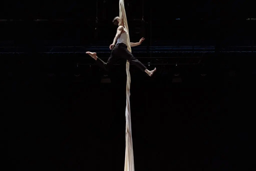 L'acrobate Antoine Boissereau se produit sur des soies aériennes blanches, suspendu en l'air, les jambes tendues et les bras agrippés au tissu, sur un fond sombre dramatique.