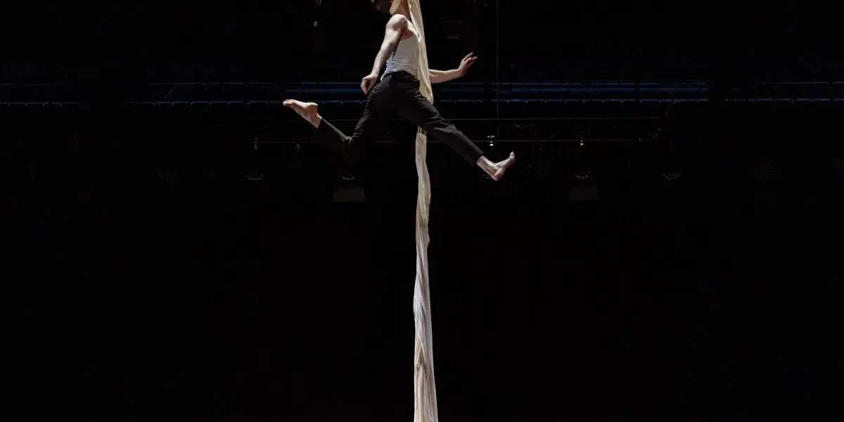 L'acrobate Antoine Boissereau se produit sur des soies aériennes blanches, suspendu en l'air, les jambes tendues et les bras agrippés au tissu, sur un fond sombre dramatique.