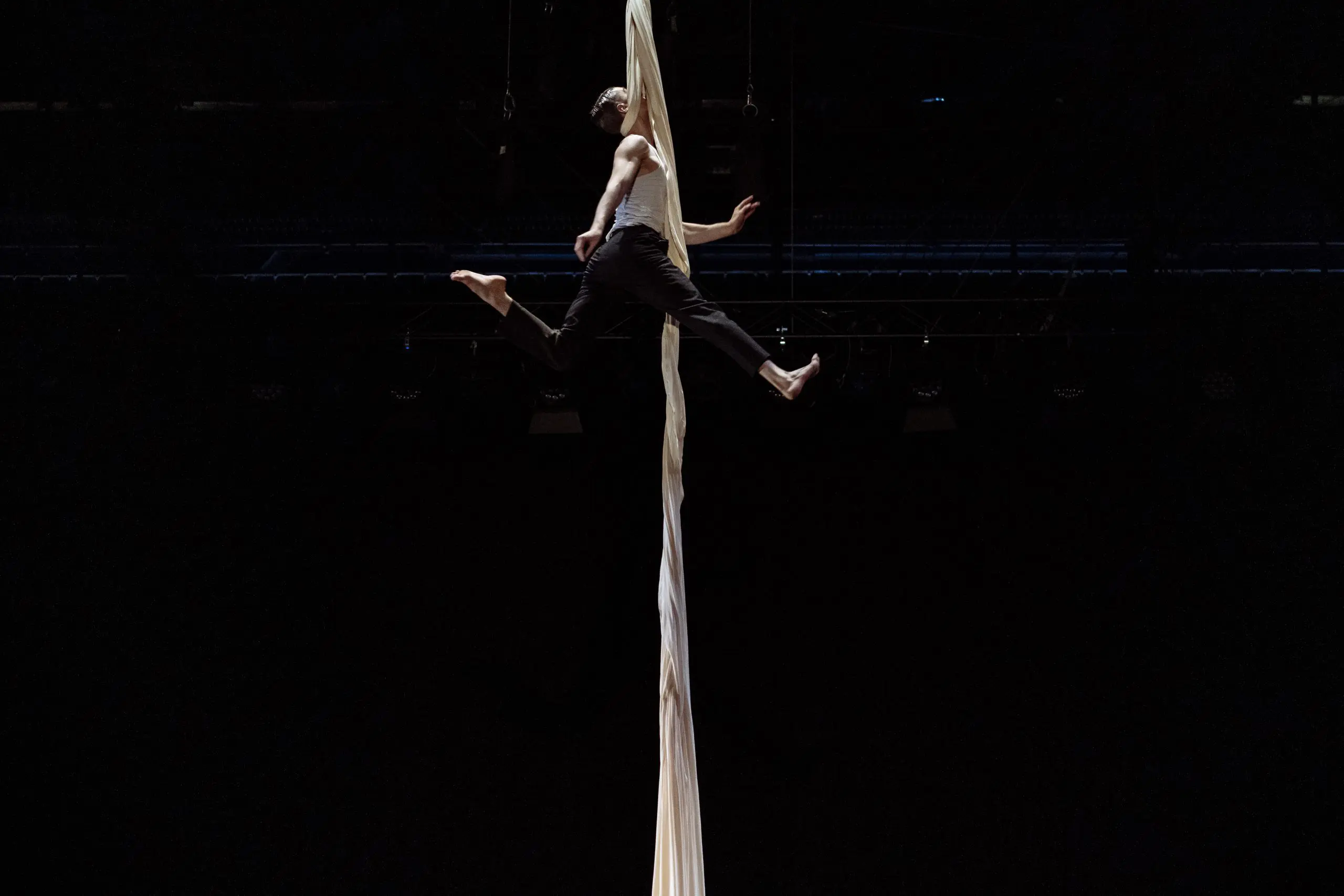 L'acrobate Antoine Boissereau se produit sur des soies aériennes blanches, suspendu en l'air, les jambes tendues et les bras agrippés au tissu, sur un fond sombre dramatique.