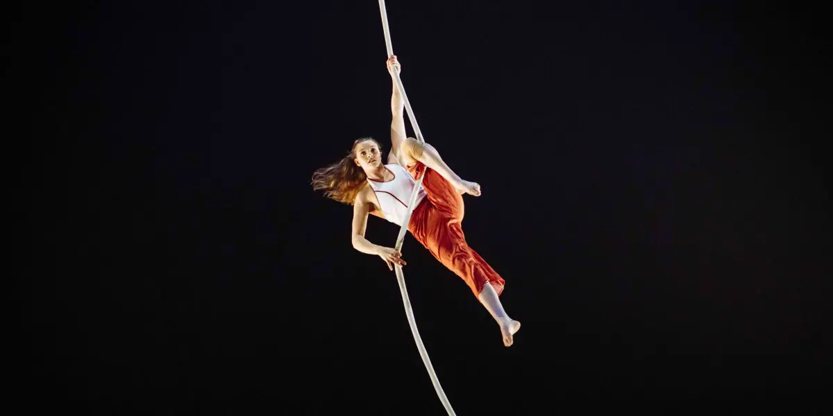 L'artiste aérienne Rosaleen Rogmans, vêtue d'un haut blanc et d'un pantalon rouge, se suspend gracieusement à une corde blanche sur un fond sombre, en équilibre avec une jambe crochetée et les cheveux flottant au milieu du mouvement.