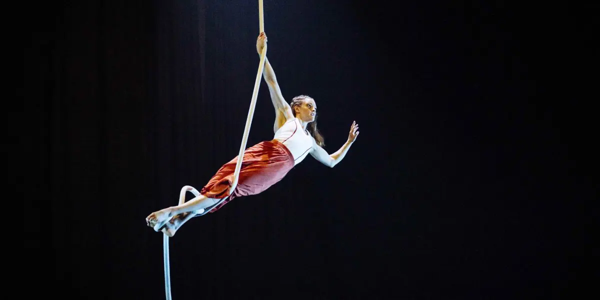 L'artiste aérienne Saffi Watson, vêtue d'un haut blanc et d'une jupe rouge, se suspend gracieusement à une corde, un bras tendu, sur un fond de scène sombre, tandis qu'un projecteur l'éclaire.