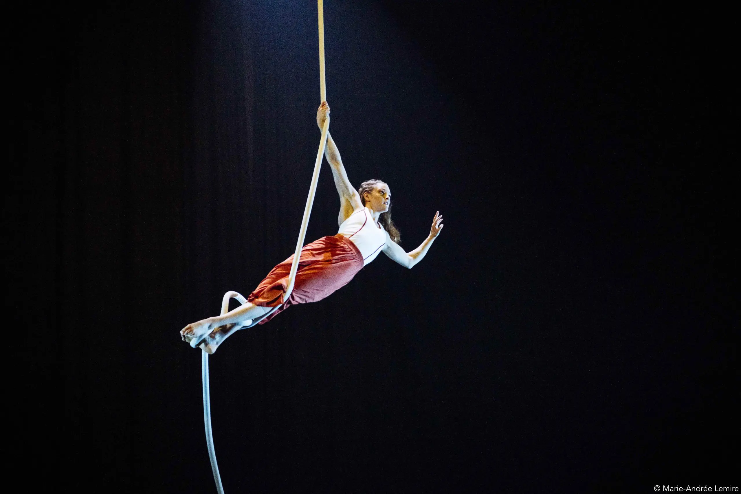 L'artiste aérienne Saffi Watson, vêtue d'un haut blanc et d'une jupe rouge, se suspend gracieusement à une corde, un bras tendu, sur un fond de scène sombre, tandis qu'un projecteur l'éclaire.