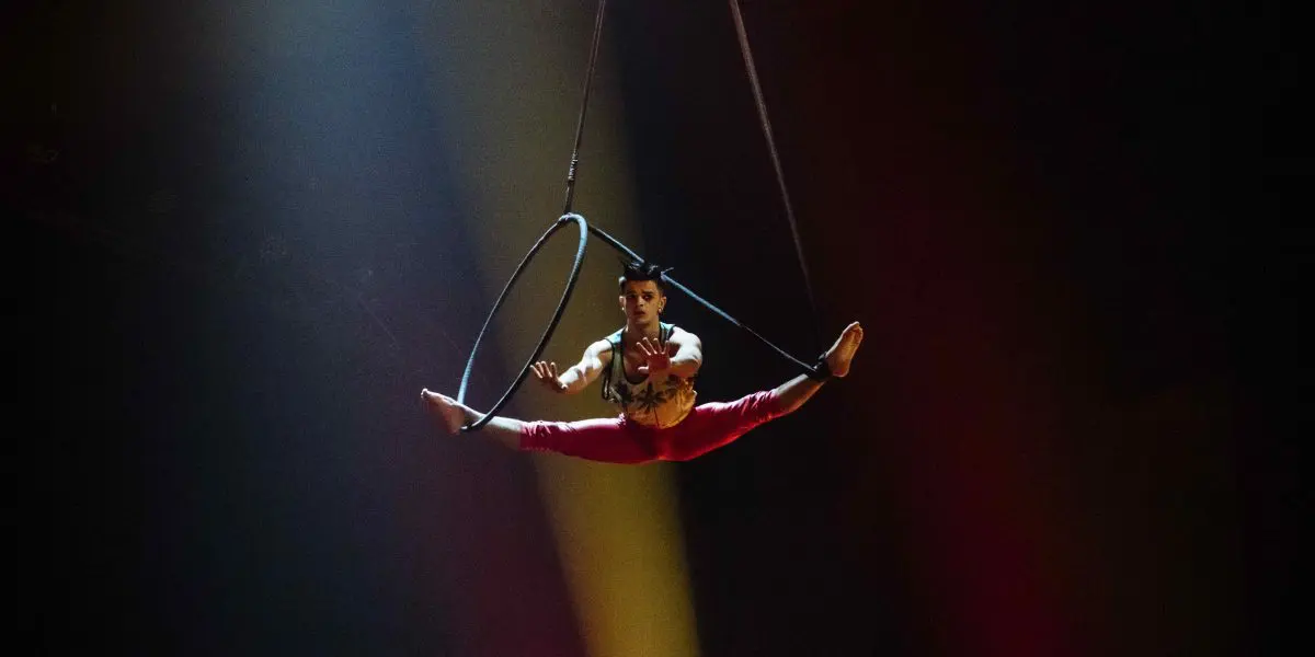 Un artiste de cirque en pantalon rouge se tient en équilibre sur un cerceau aérien, suspendu dans les airs sous un éclairage de scène dramatique sur fond sombre, incarnant l'art d'Enzo Pebre.