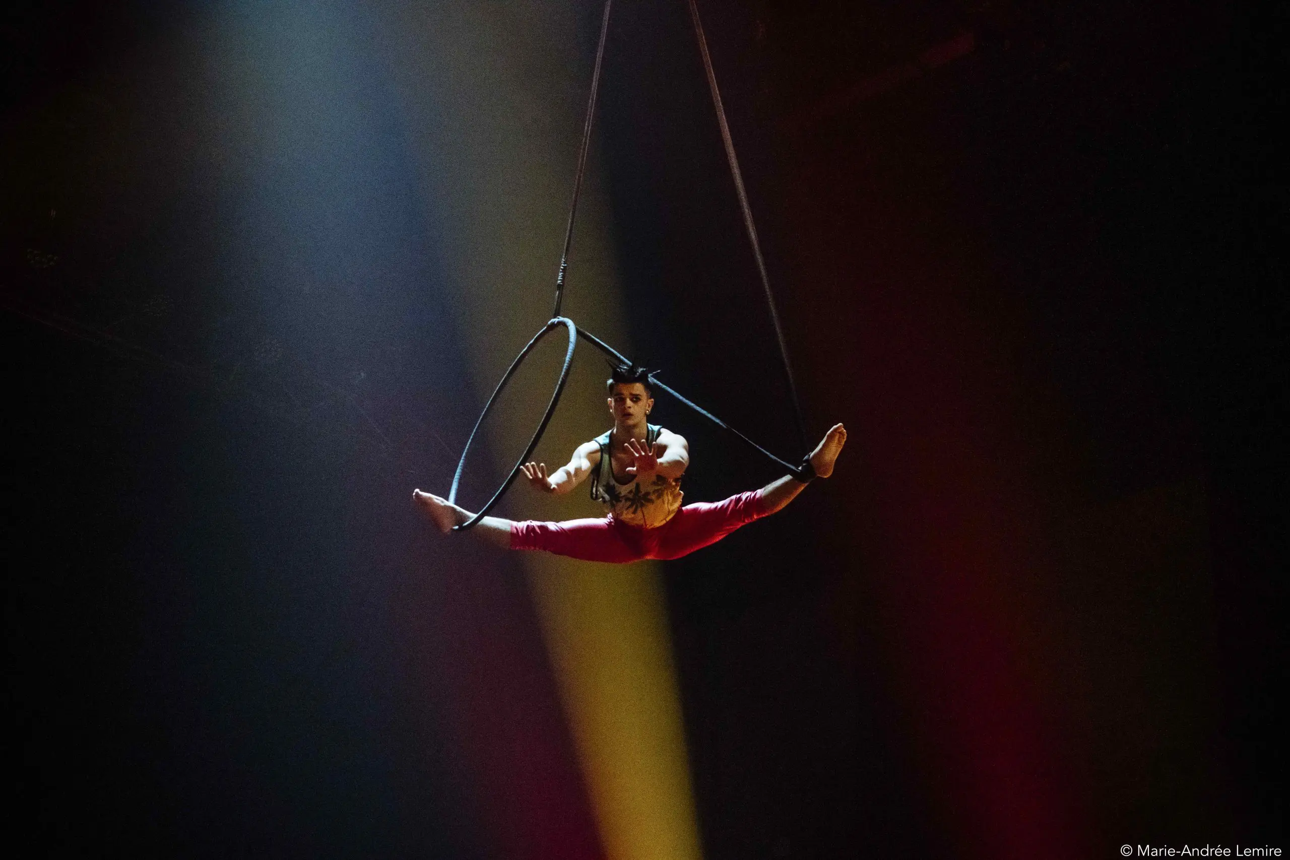 Un artiste de cirque en pantalon rouge se tient en équilibre sur un cerceau aérien, suspendu dans les airs sous un éclairage de scène dramatique sur fond sombre, incarnant l'art d'Enzo Pebre.