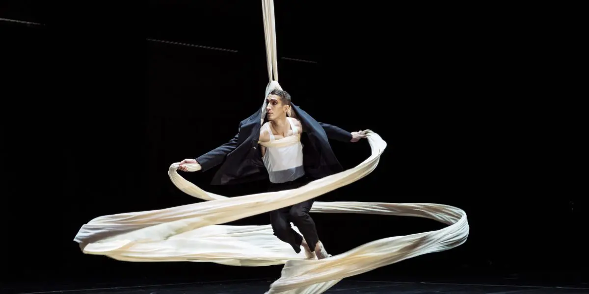 Antoine Boissereau, vêtu d'un costume noir, exécute une danse aérienne sur soie, suspendu dans les airs par un long tissu blanc qui tourbillonne dramatiquement autour de lui sur un fond sombre.