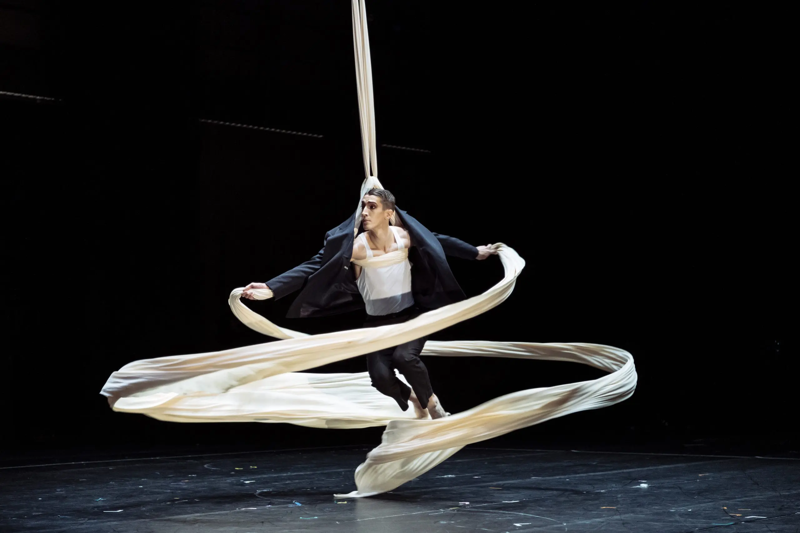Antoine Boissereau, vêtu d'un costume noir, exécute une danse aérienne sur soie, suspendu dans les airs par un long tissu blanc qui tourbillonne dramatiquement autour de lui sur un fond sombre.