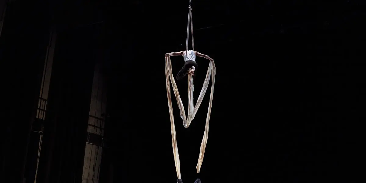 Un artiste aérien est suspendu à l'envers par de longs rubans de soie dans un théâtre sombre. Antoine Boissereau se tient sur la scène en contrebas.