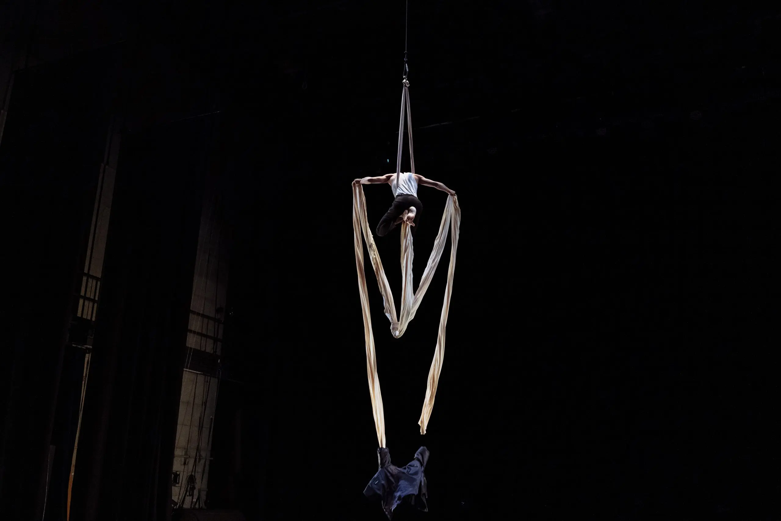 Un artiste aérien est suspendu à l'envers par de longs rubans de soie dans un théâtre sombre. Antoine Boissereau se tient sur la scène en contrebas.