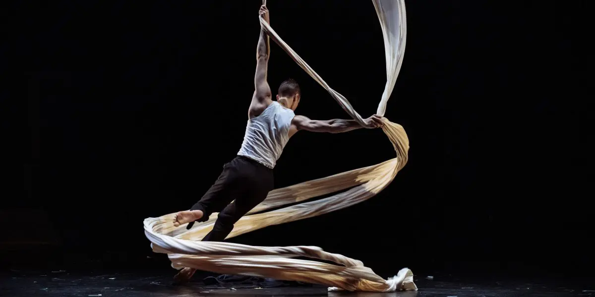 L'artiste aérien Antoine Boissereau se produit sur scène, suspendu par de longs rubans de soie blanche. Vêtu d'un débardeur blanc et d'un pantalon noir, il se tortille gracieusement dans les airs, créant des formes dynamiques avec le tissu sur un fond sombre.