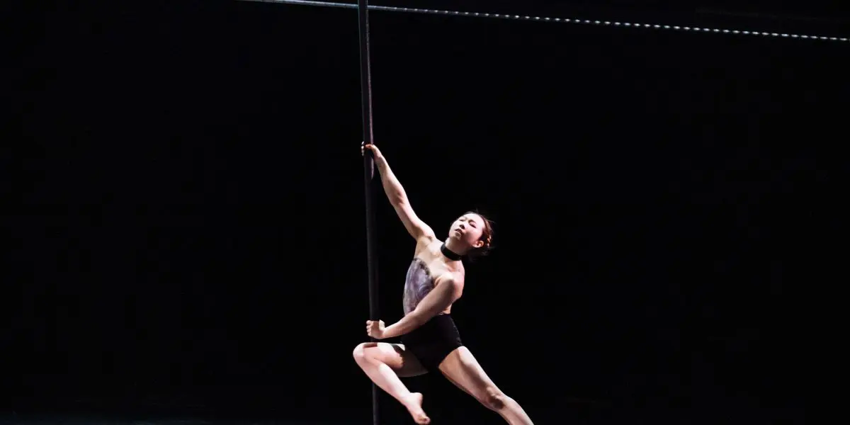 Minji Kim, vêtue de noir, exécute un mouvement gracieux de pole dance sur scène, en équilibre avec un bras et une jambe tendus sur un fond sombre.