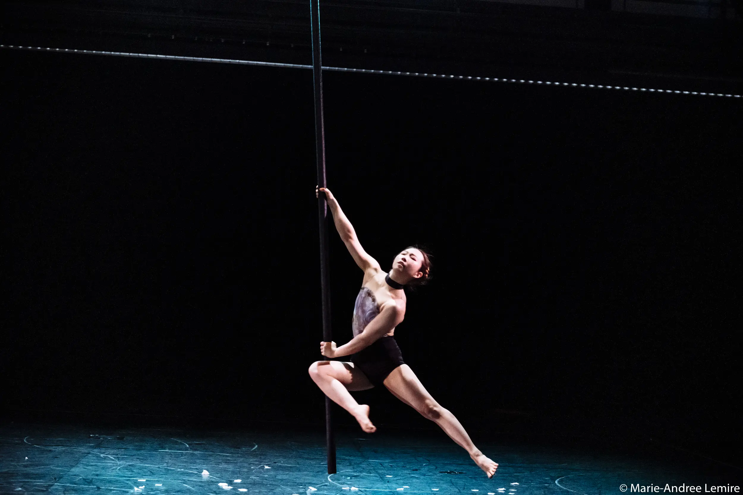 Minji Kim, vêtue de noir, exécute un mouvement gracieux de pole dance sur scène, en équilibre avec un bras et une jambe tendus sur un fond sombre.