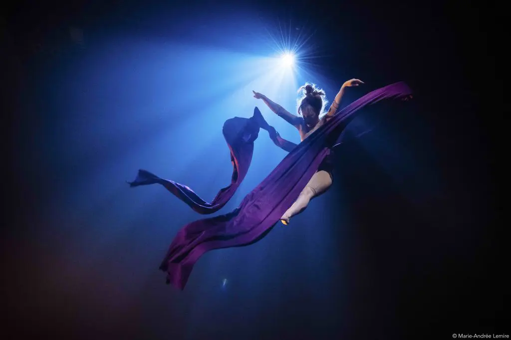 Une artiste aérienne, Margaux Wasteels, est suspendue dans les airs, gracieusement enveloppée dans un tissu violet fluide, tandis qu'un éclairage de scène dramatique crée un effet de lumière derrière elle.