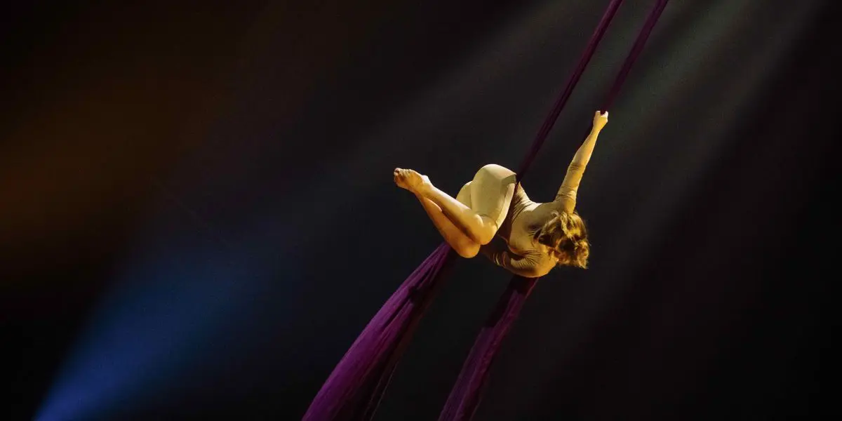 L'artiste aérienne Margaux Wasteels, dans un costume beige, est suspendue dans les airs, posant gracieusement sur des soies violettes profondes, un projecteur la mettant en valeur sur un fond sombre.