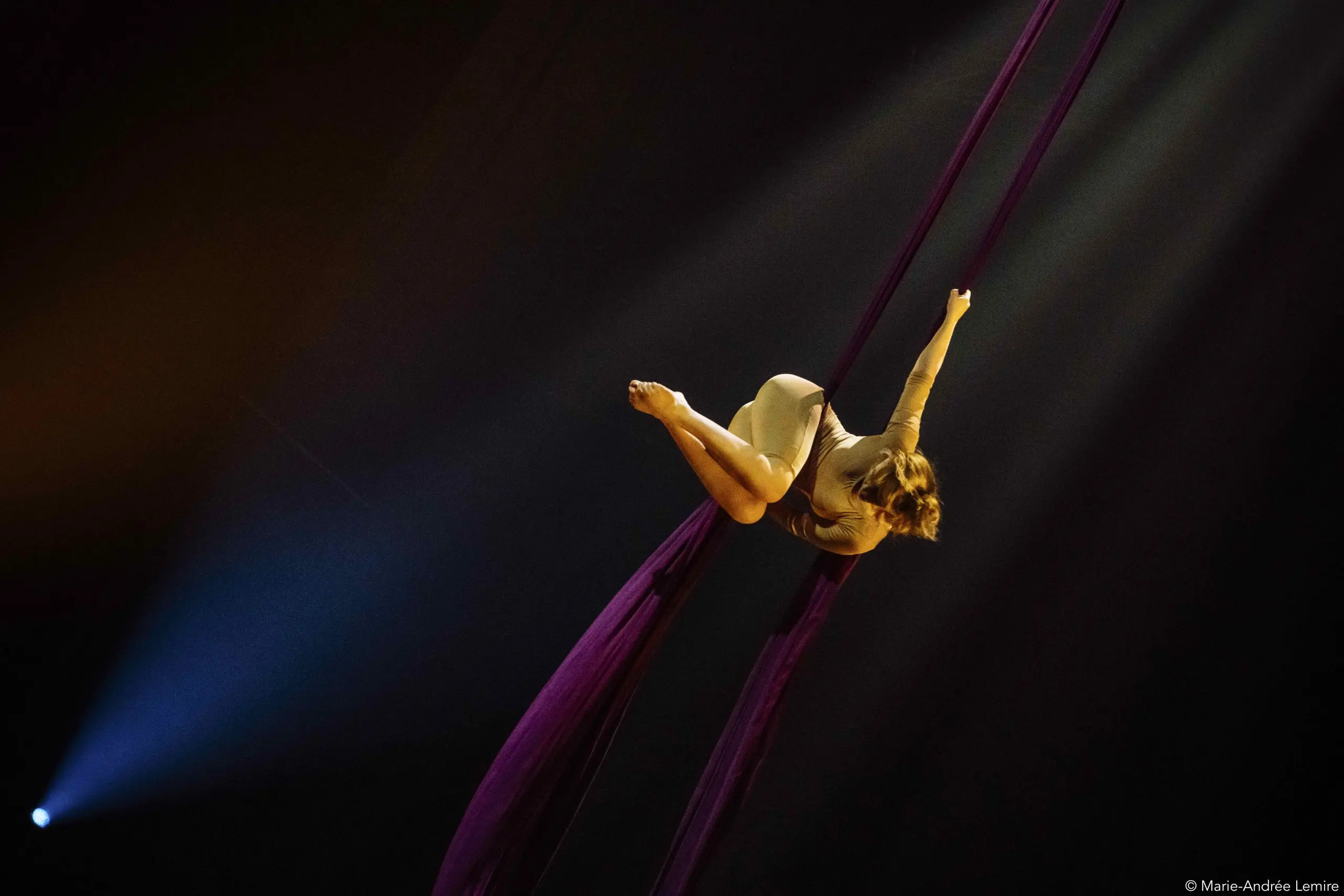 L'artiste aérienne Margaux Wasteels, dans un costume beige, est suspendue dans les airs, posant gracieusement sur des soies violettes profondes, un projecteur la mettant en valeur sur un fond sombre.