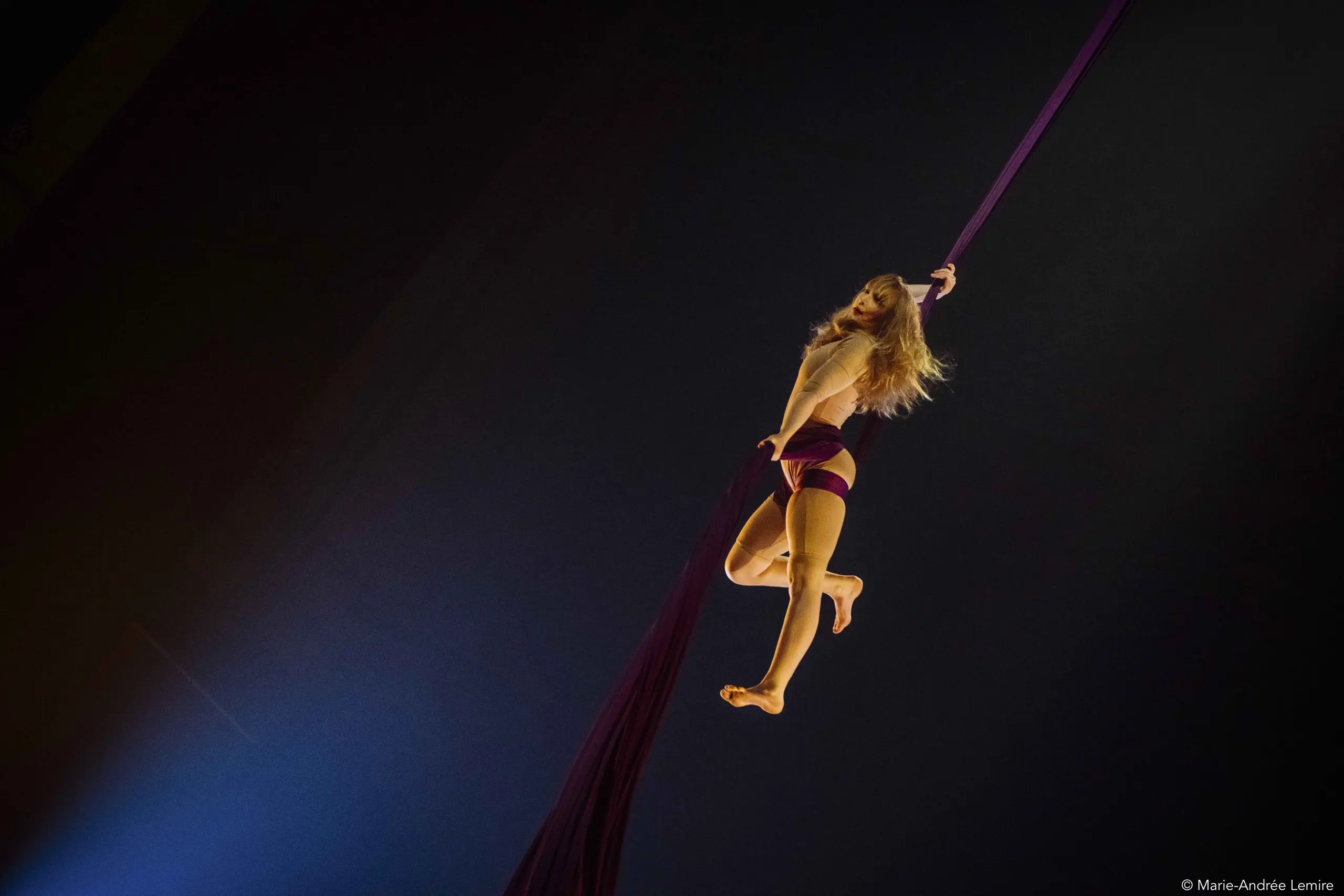 L'artiste aérienne Margaux Wasteels est suspendue dans les airs, agrippant des soies violettes d'une main et d'une jambe tendue, éclairée par la lumière de la scène sur un fond sombre.