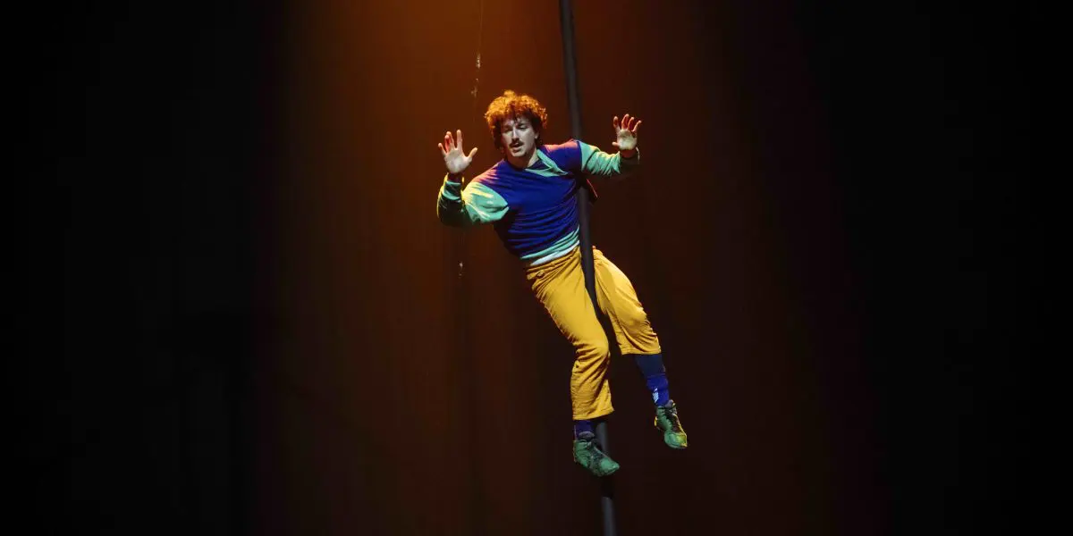 Un artiste de cirque aux vêtements colorés s'accroche à un poteau vertical, suspendu dans les airs sous un projecteur sur un fond sombre, mettant en valeur l'art captivant de Simon Lemire.