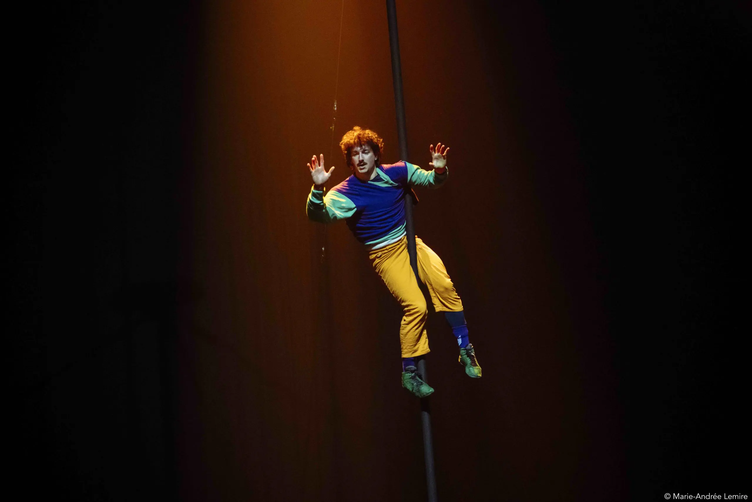 Un artiste de cirque aux vêtements colorés s'accroche à un poteau vertical, suspendu dans les airs sous un projecteur sur un fond sombre, mettant en valeur l'art captivant de Simon Lemire.