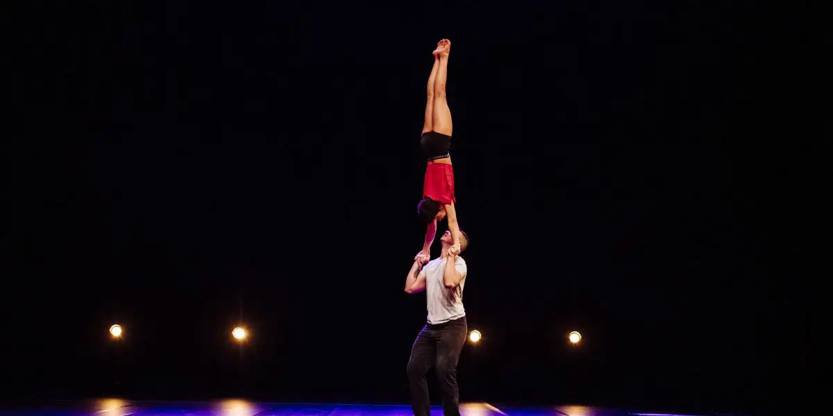 Deux acrobates, Michael Carter et Frida Velasco, se produisent sur scène ; l'un se tient debout et soulève l'autre verticalement par les jambes au-dessus de sa tête. L'arrière-plan est sombre, avec quatre projecteurs.