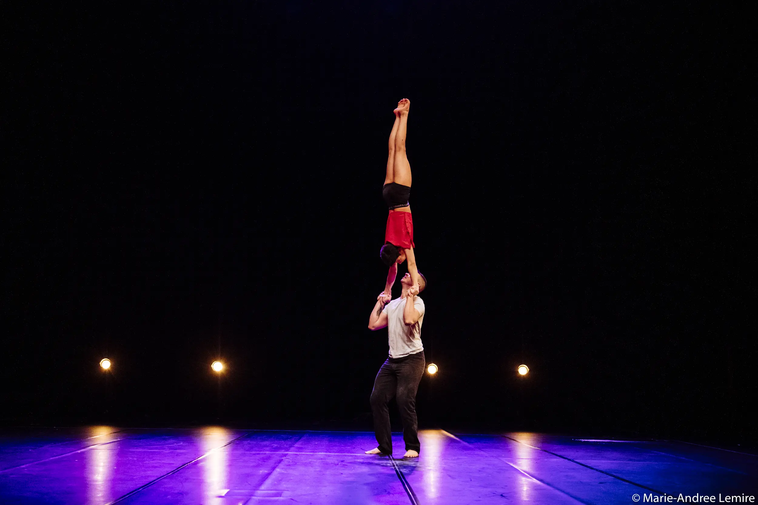 Deux acrobates, Michael Carter et Frida Velasco, se produisent sur scène ; l'un se tient debout et soulève l'autre verticalement par les jambes au-dessus de sa tête. L'arrière-plan est sombre, avec quatre projecteurs.