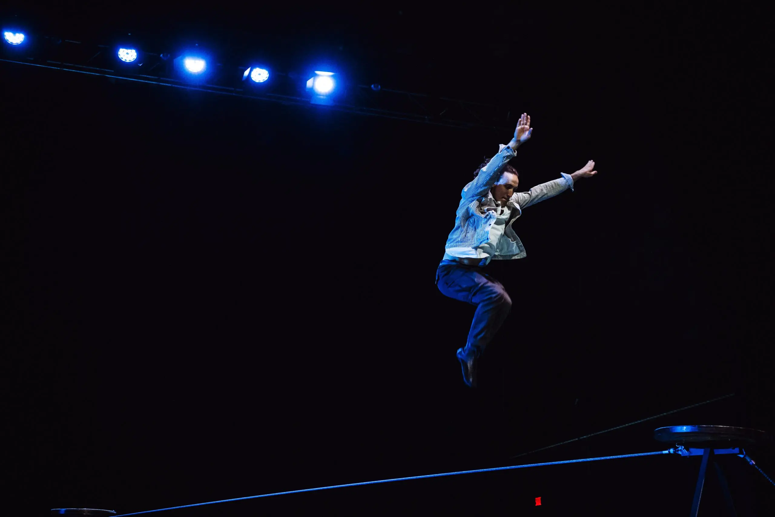 Une personne en costume saute les bras levés au-dessus d'une corde raide sur une scène sombre, éclairée par des projecteurs bleus depuis le haut, capturant l'esprit audacieux de Joel Malkoff.