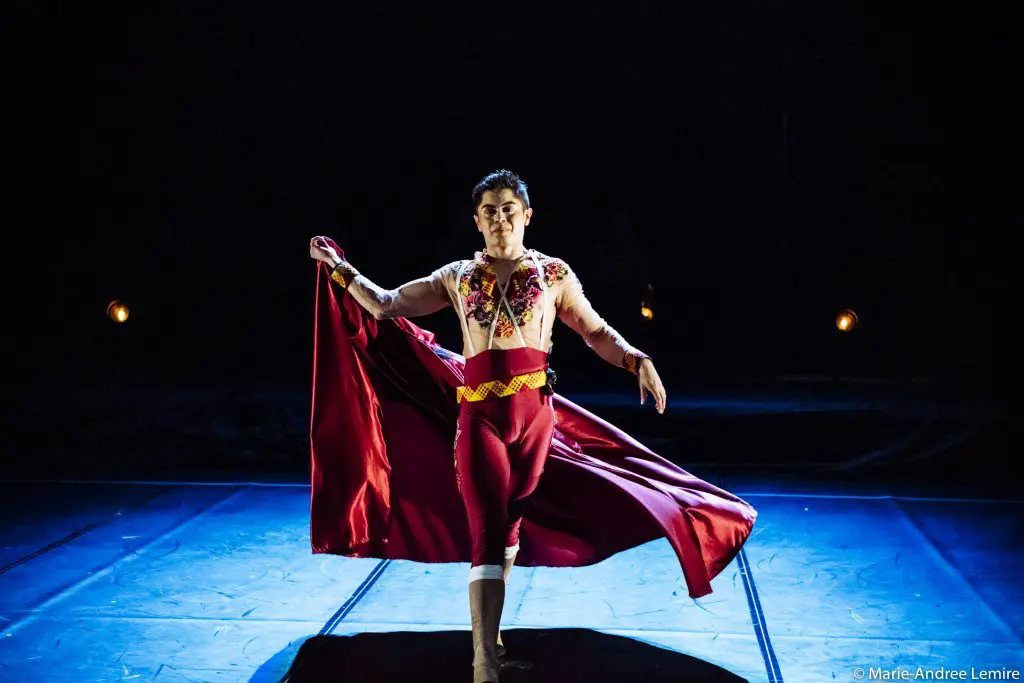 Gabriel Olmos Cruz, un danseur de ballet vêtu d'un costume inspiré des matadors, pose gracieusement sur scène, tenant un pan d'une cape rouge fluide, sous un éclairage dramatique et un arrière-plan sombre.