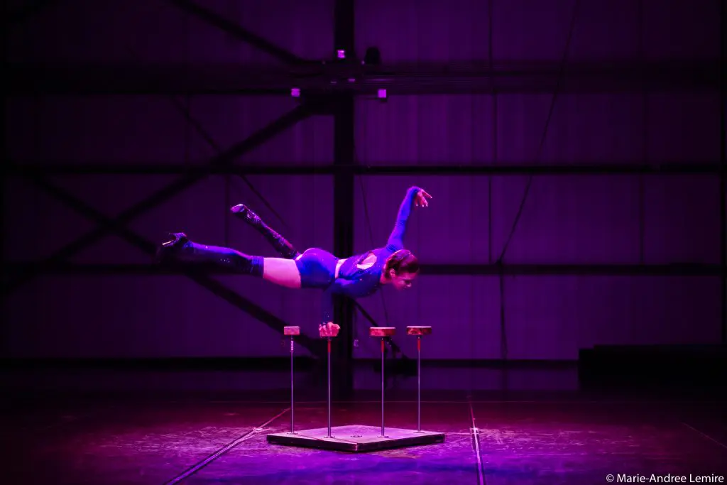Une acrobate en tenue bleue se tient en équilibre sur ses mains au sommet de cannes, étendant ses jambes vers l'arrière dans un mouvement de fente. La scène, éclairée par des lumières violettes et conçue par Yann Lortie, comporte une grande structure métallique en arrière-plan.