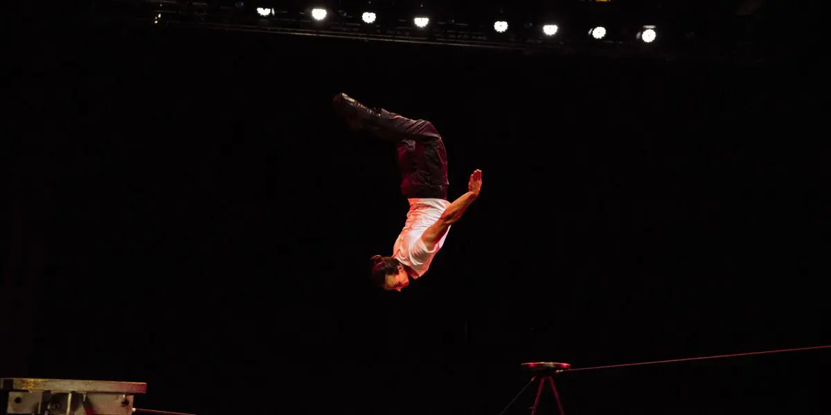 Joel Malkoff, cheveux longs en queue de cheval, vêtu d'une chemise blanche et d'un pantalon sombre, effectue un saut périlleux au-dessus d'une corde raide sur une scène sombre éclairée par les lumières de la scène.