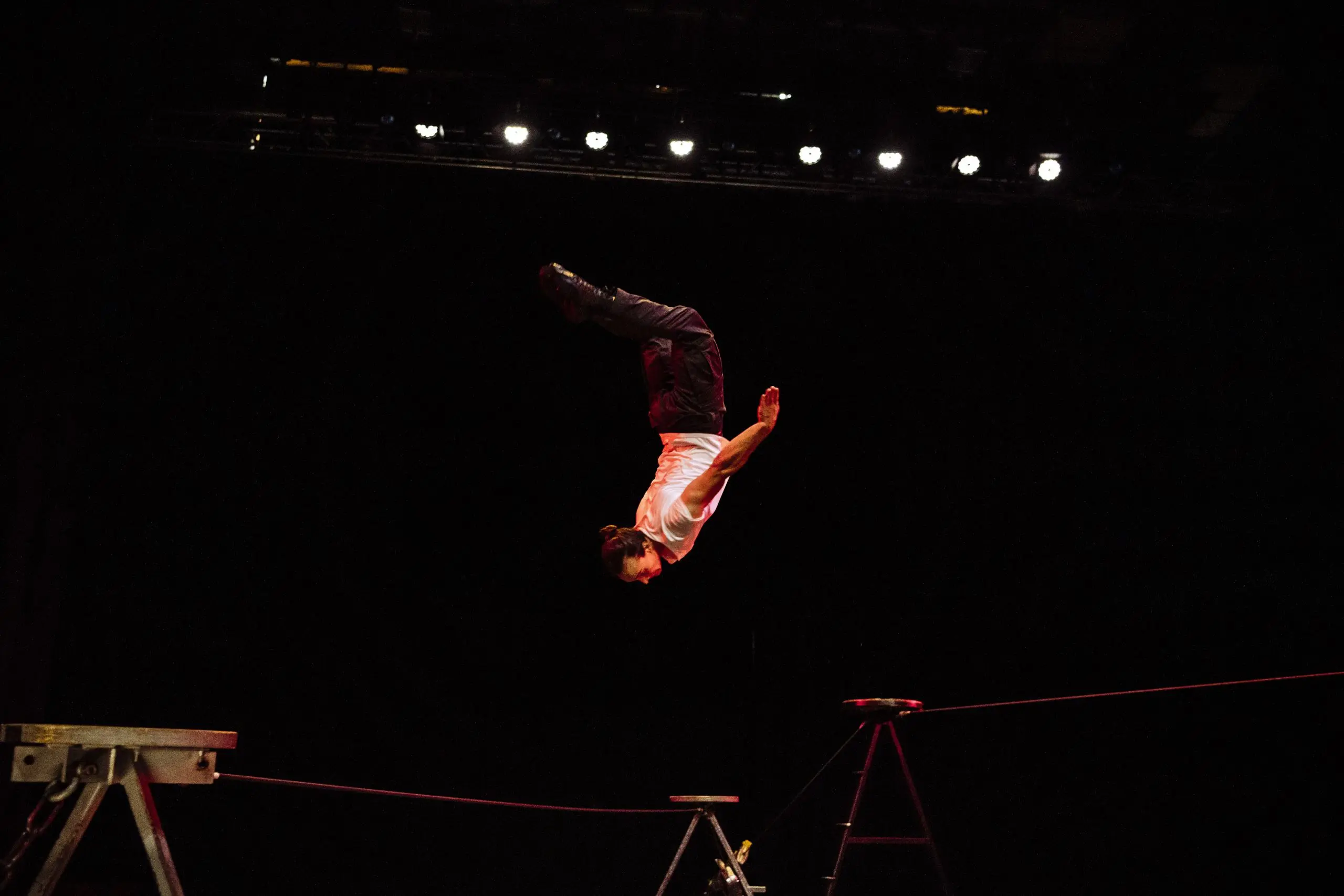 Joel Malkoff, cheveux longs en queue de cheval, vêtu d'une chemise blanche et d'un pantalon sombre, effectue un saut périlleux au-dessus d'une corde raide sur une scène sombre éclairée par les lumières de la scène.