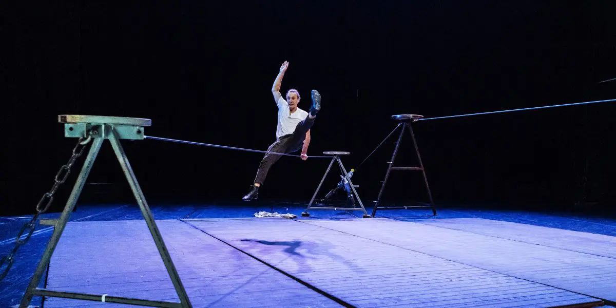 Joel Malkoff, un artiste vêtu d'une chemise blanche et d'un pantalon sombre, se tient en équilibre, une jambe levée, assis sur une slackline, suspendue entre deux supports métalliques sur une scène à l'éclairage bleu.