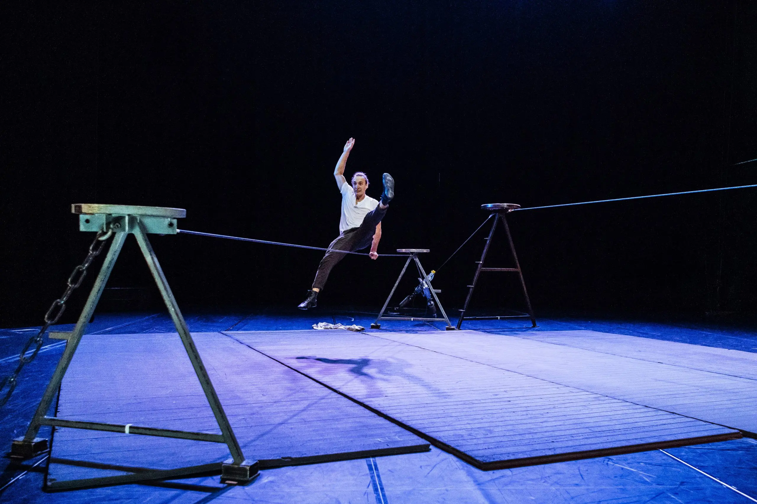 Joel Malkoff, un artiste vêtu d'une chemise blanche et d'un pantalon sombre, se tient en équilibre, une jambe levée, assis sur une slackline, suspendue entre deux supports métalliques sur une scène à l'éclairage bleu.