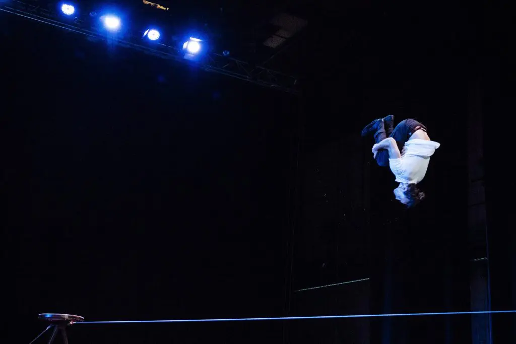 Joel Malkoff, vêtu d'une chemise blanche et d'un pantalon sombre, effectue un saut périlleux arrière au-dessus d'une corde raide sous les lumières bleues de la scène, un tabouret étant visible en dessous, à gauche.