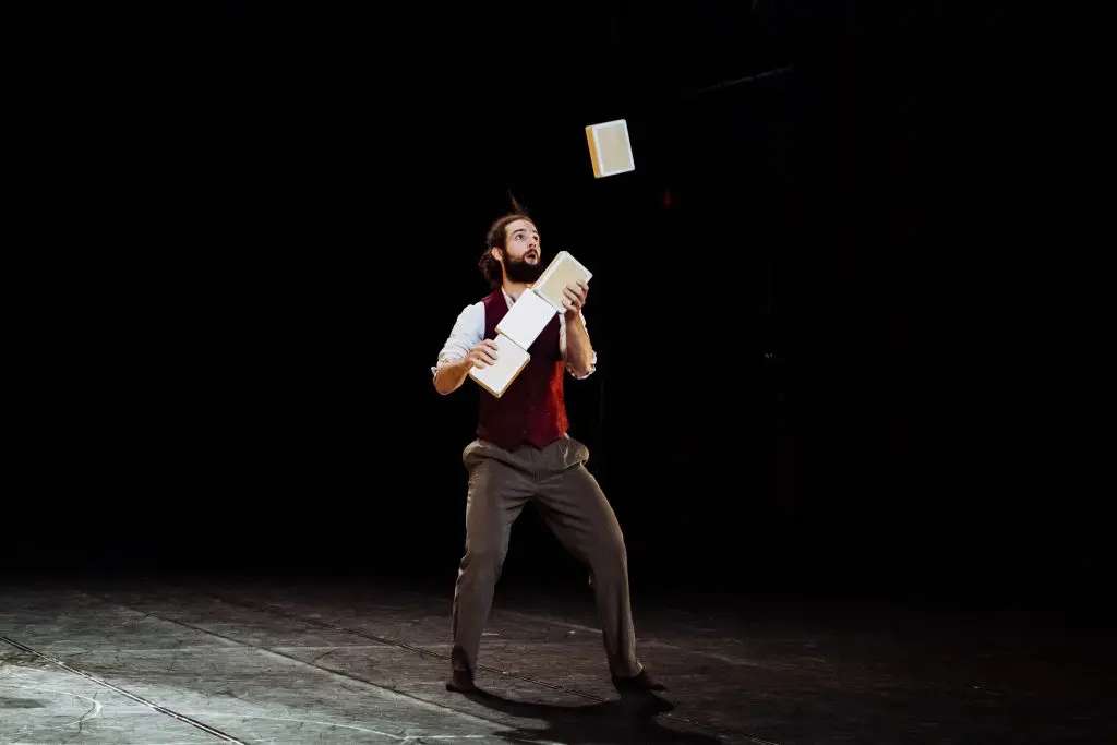 Joaquim Verrier, vêtu d'un gilet rouge et d'un pantalon habillé, jongle avec trois livres sur une scène faiblement éclairée, entièrement concentré sur sa performance.