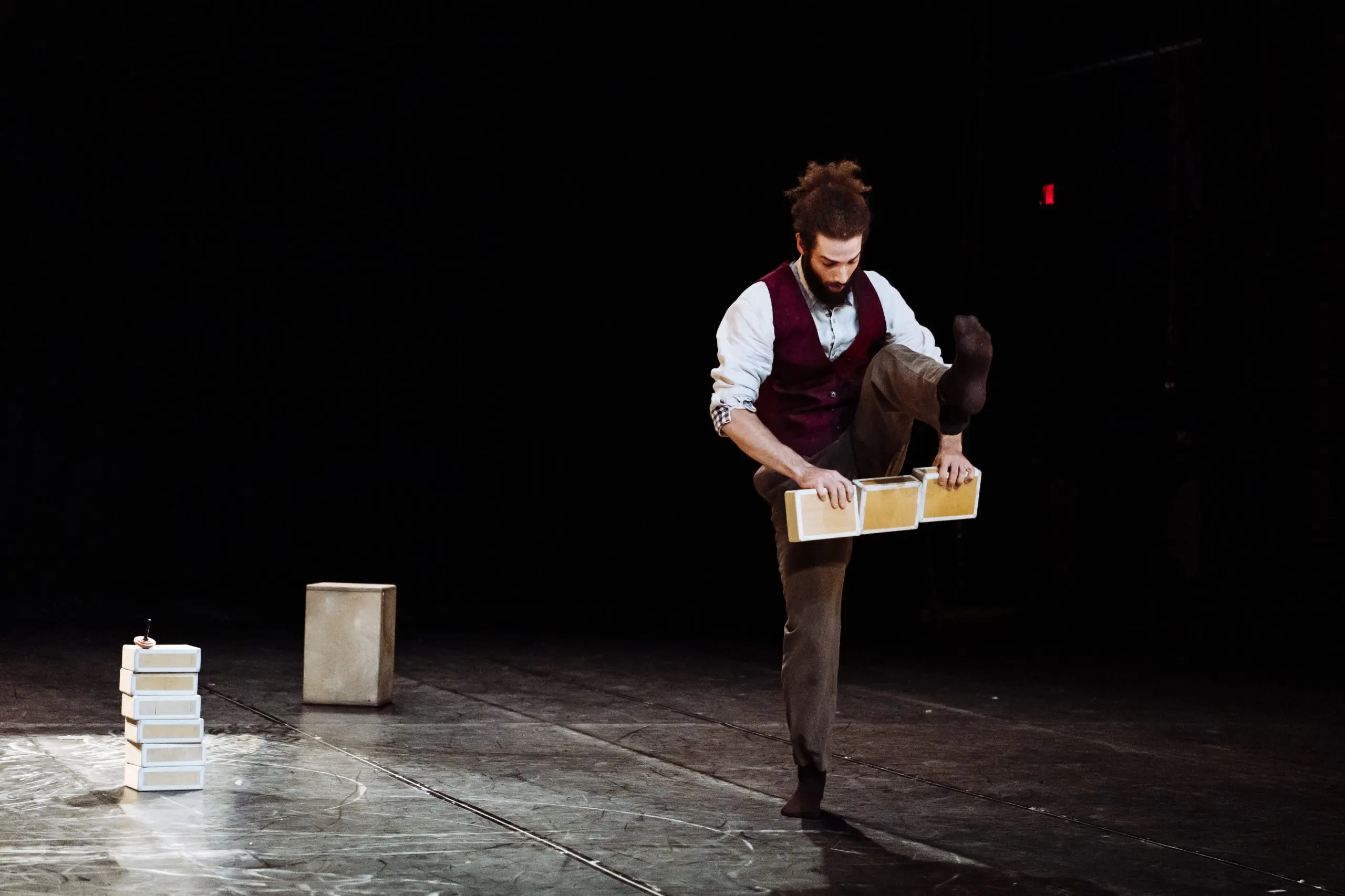 Joaquim Verrier, un artiste barbu aux cheveux attachés, se tient en équilibre sur une jambe tout en tenant des blocs de bois sur scène. D'autres blocs et un cube sont empilés à l'arrière-plan sur une toile de fond sombre.