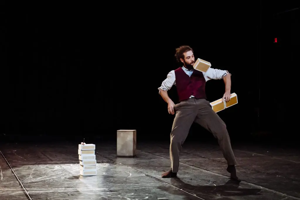 Un artiste pieds nus, Joaquim Verrier, vêtu d'un gilet et d'un pantalon habillé, équilibre et jongle avec trois objets rectangulaires beiges sur scène, en tenant l'un d'eux dans sa bouche, avec une petite pile et un cube à l'arrière-plan sur une toile de fond sombre.