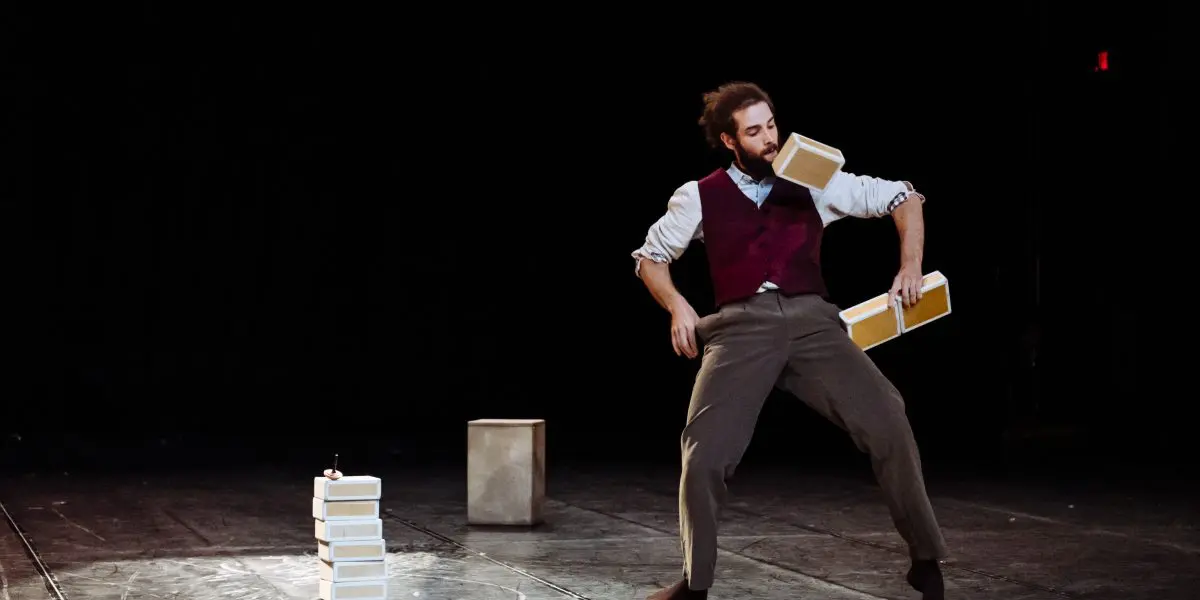 Un artiste pieds nus, Joaquim Verrier, vêtu d'un gilet et d'un pantalon habillé, équilibre et jongle avec trois objets rectangulaires beiges sur scène, en tenant l'un d'eux dans sa bouche, avec une petite pile et un cube à l'arrière-plan sur une toile de fond sombre.