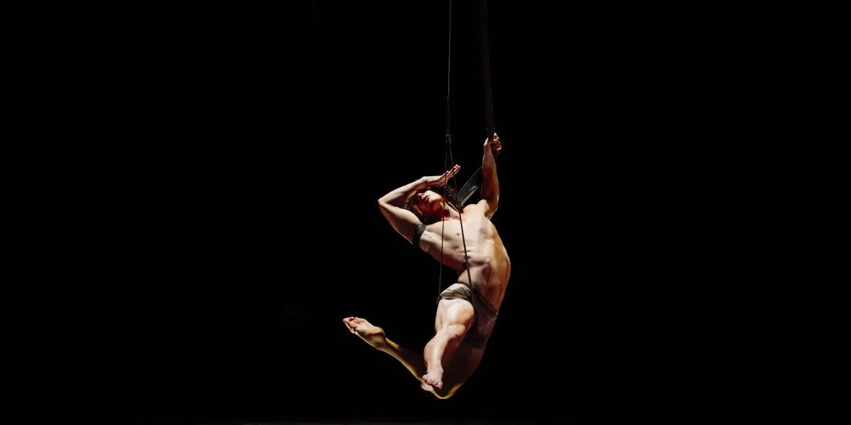 L'artiste aérien Agustin Rodriguez Beltran se produit suspendu à des cordes sur un fond sombre, le corps gracieusement arqué, un bras au-dessus de la tête et une jambe pliée, éclairé par un projecteur.