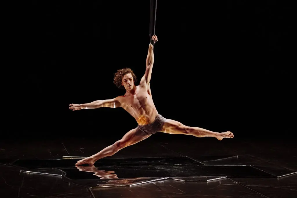 Un artiste aérien, Agustin Rodriguez Beltran, vêtu d'un simple slip, se suspend d'une seule main à une sangle et étend ses jambes en position fendue au-dessus d'une scène sombre et réfléchissante.