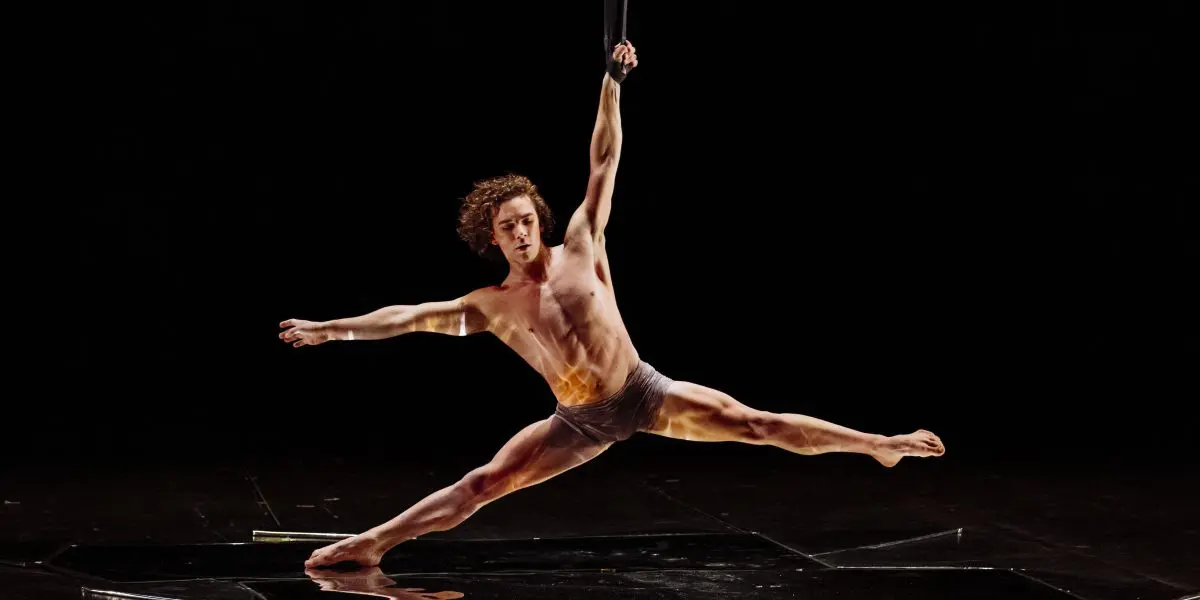 Un artiste aérien, Agustin Rodriguez Beltran, vêtu d'un simple slip, se suspend d'une seule main à une sangle et étend ses jambes en position fendue au-dessus d'une scène sombre et réfléchissante.
