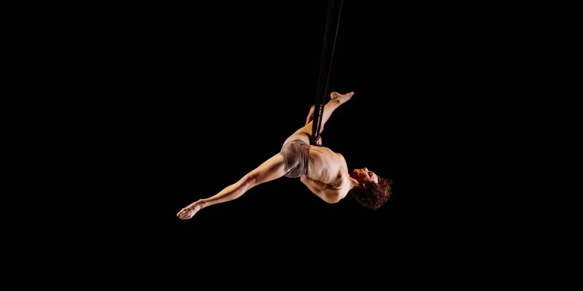 L'artiste aérien Agustin Rodriguez Beltran est suspendu dans les airs par des sangles noires sur un fond noir, en équilibre horizontal avec une jambe tendue et l'autre pliée, faisant preuve d'une force et d'une souplesse impressionnantes.