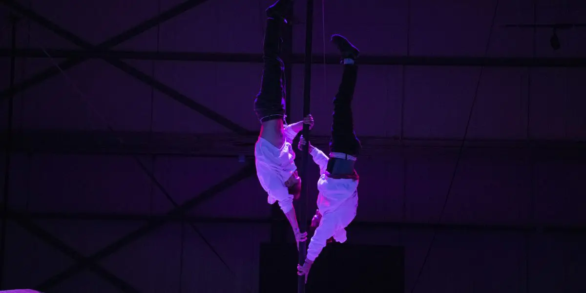 Deux acrobates vêtus de chemises blanches, dont Mathys Guyon Matte, sont suspendus à un poteau vertical, se tenant par la main, sur un fond industriel faiblement éclairé par des lumières violettes.