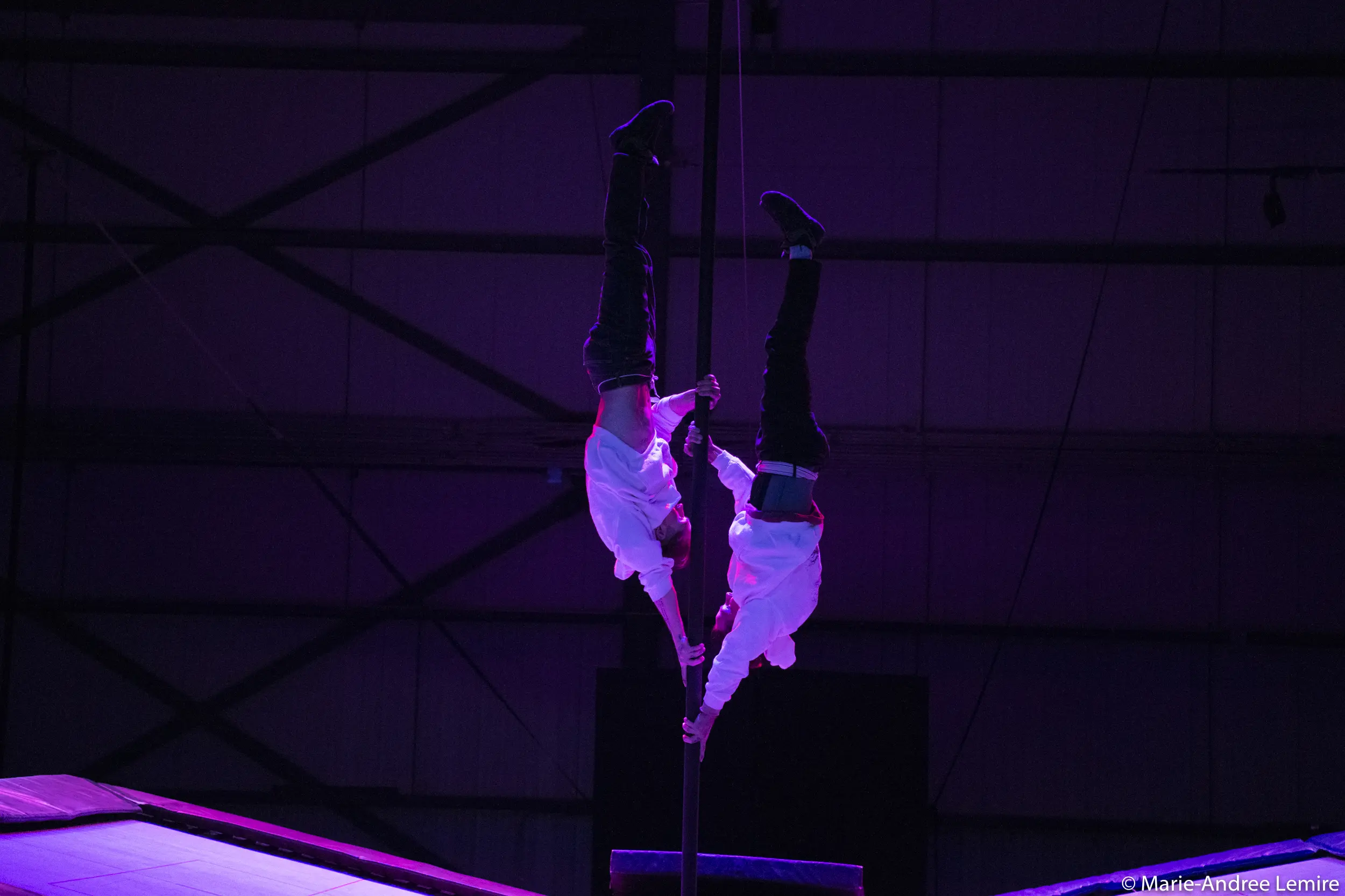 Deux acrobates vêtus de chemises blanches, dont Mathys Guyon Matte, sont suspendus à un poteau vertical, se tenant par la main, sur un fond industriel faiblement éclairé par des lumières violettes.