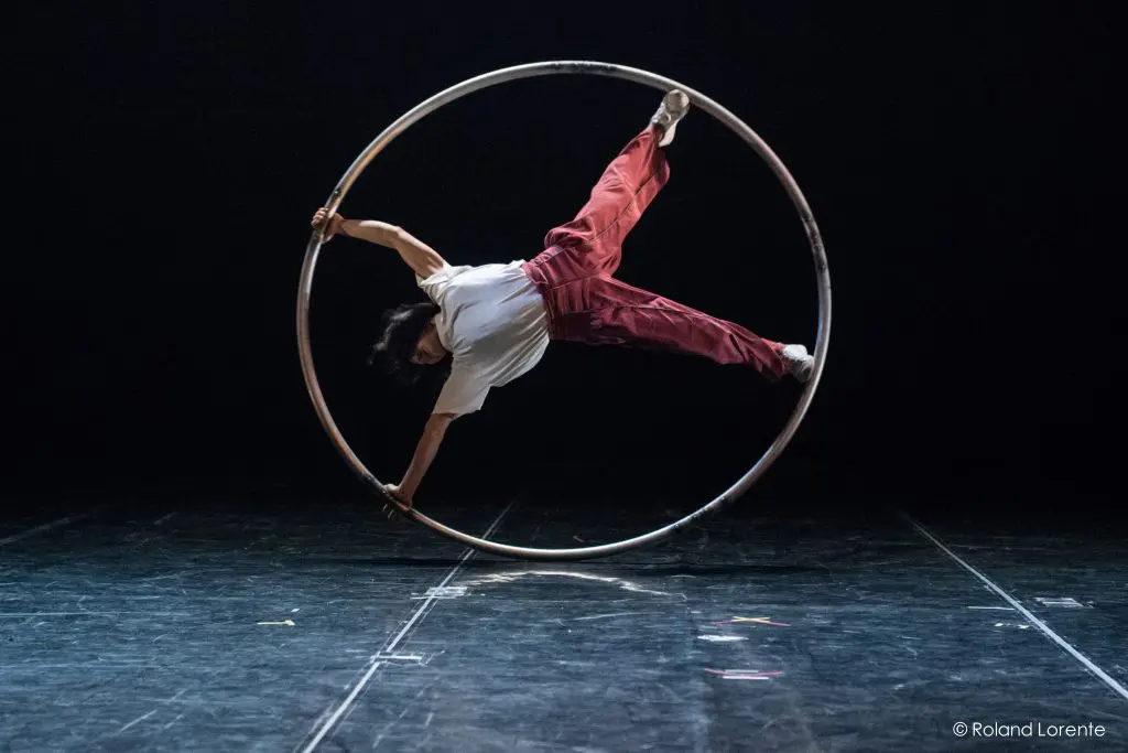 Miho Inaba, vêtue d'une chemise blanche et d'un pantalon rouge, se tient en équilibre horizontal à l'intérieur d'une grande roue métallique sur scène. Avec une main et un pied qui touchent la roue, elle se produit sur un fond sombre.