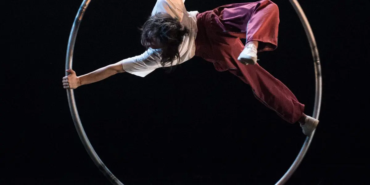 Miho Inaba se tient habilement en équilibre à l'intérieur d'un grand cerceau métallique, exécutant un mouvement acrobatique sur une scène sombre. Ils sont vêtus d'une chemise blanche et d'un pantalon ample de couleur bordeaux, une main et un pied agrippés au cerceau.