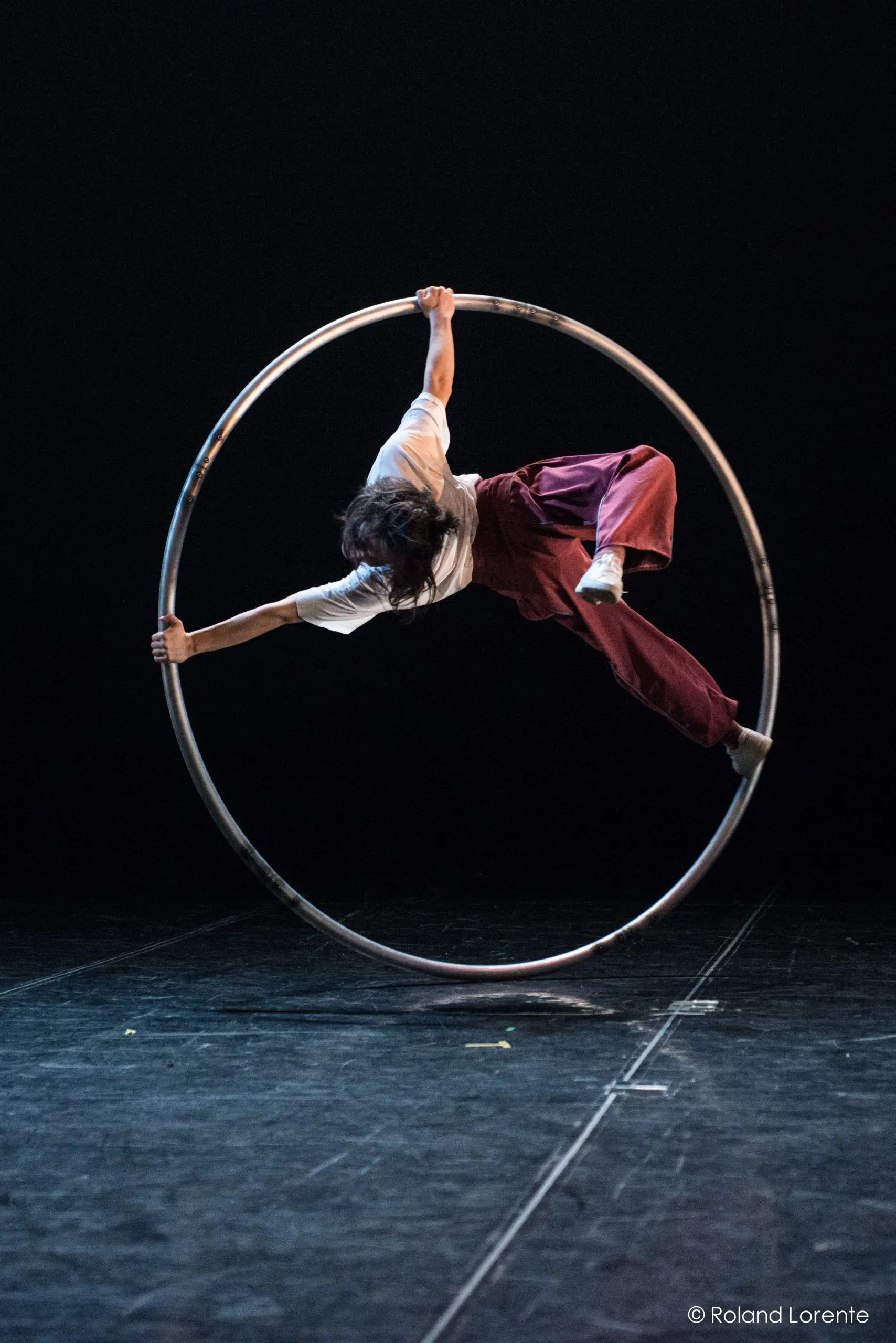 Miho Inaba se tient habilement en équilibre à l'intérieur d'un grand cerceau métallique, exécutant un mouvement acrobatique sur une scène sombre. Ils sont vêtus d'une chemise blanche et d'un pantalon ample de couleur bordeaux, une main et un pied agrippés au cerceau.